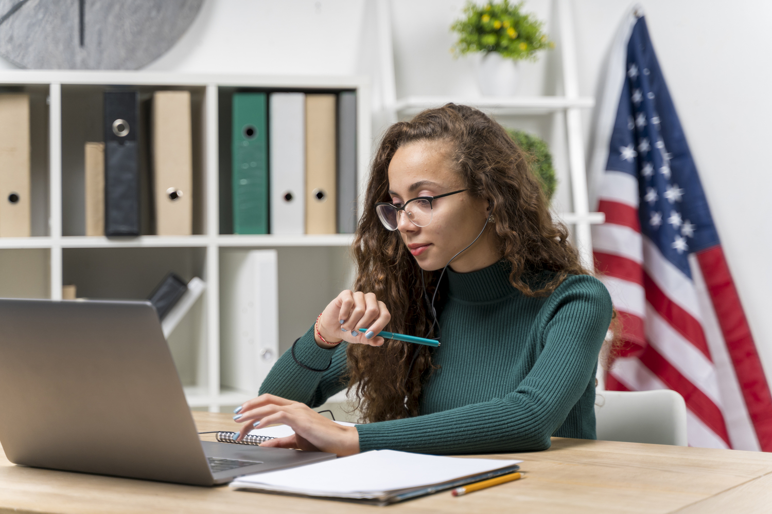 Mulher adulta estudando inglês online em ambiente profissional, usando notebook e fones de ouvido, com bandeira dos Estados Unidos ao fundo.