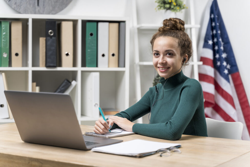 Profissional estudando inglês corporativo online em frente ao notebook, em ambiente de escritório com bandeira dos EUA ao fundo.