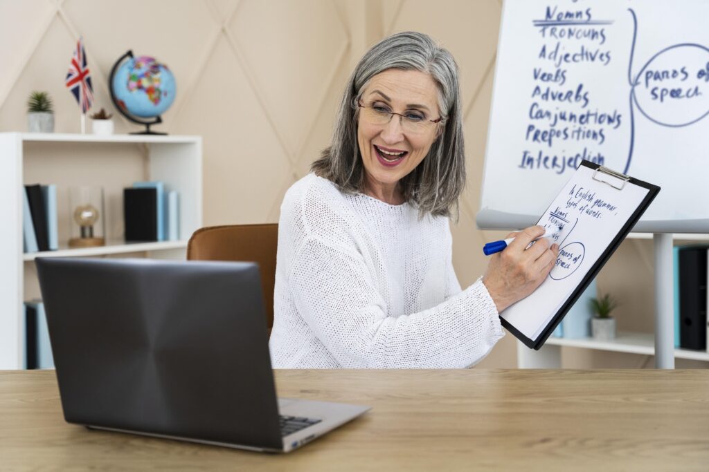 Professora ensinando inglês para adultos iniciantes em aula online, explicando gramática básica em quadro branco